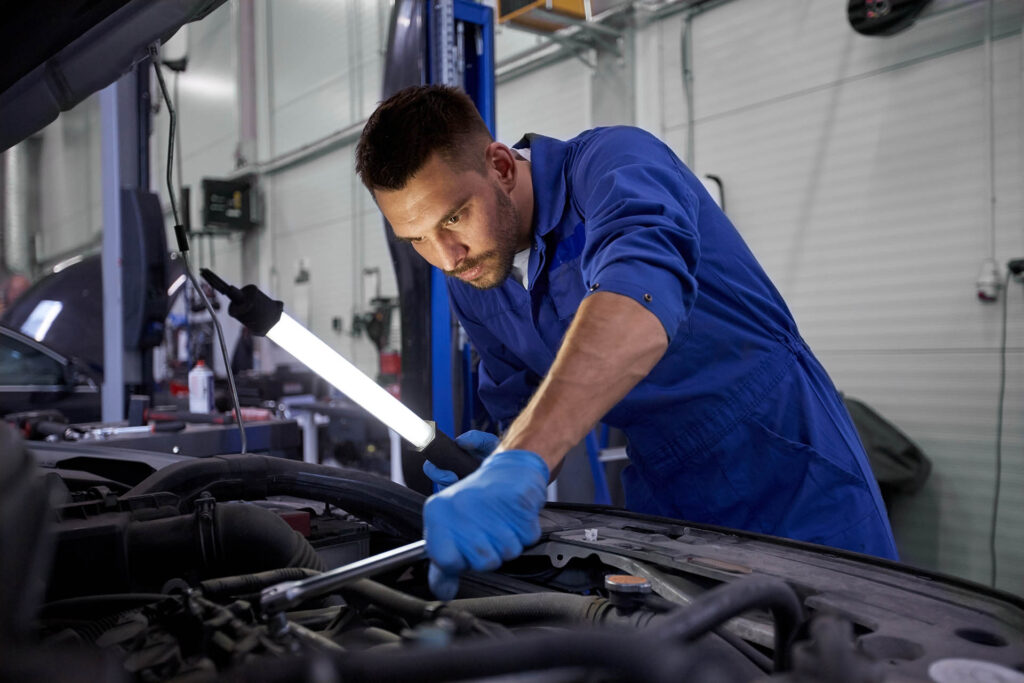 car service, repair, maintenance and people concept - auto mechanic man with lamp working at workshop